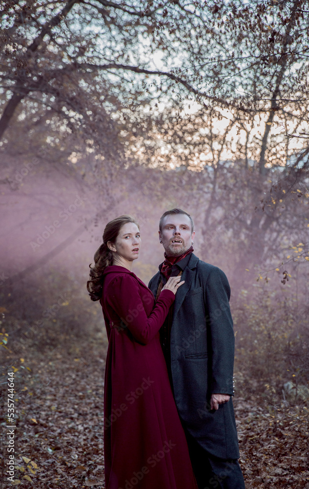 Vampire man wearing victorian suit with vampire woman in the autumn forest in the fog. Vintage medieval concept style. White and black photo.