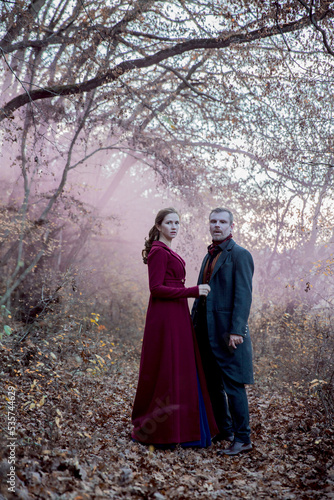 Vampire man wearing victorian suit with vampire woman in the autumn forest in the fog. Vintage medieval concept style. White and black photo.