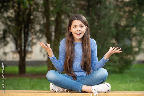 Foto Outraged girl teenager shrugging shoulders sitting legs crossed on bench outdoor