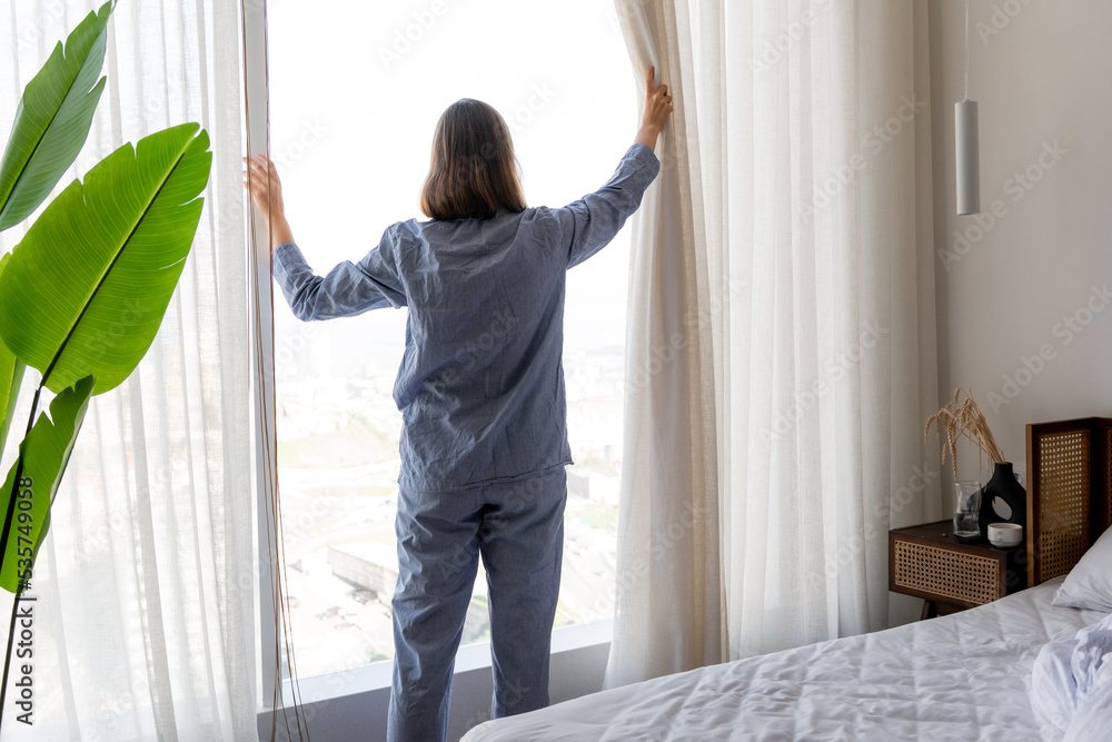 Young woman in pajamas open white curtains at the window, the morning after waking up in the bedroom of her home Young woman in pajamas open white curtains at the window, the morning after waking up in the bedroom of her home