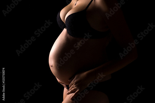 Belly of a pregnant girl in black lingerie on a black background. Beautiful pregnant belly in black lingerie. Studio shooting. Woman holding her belly.