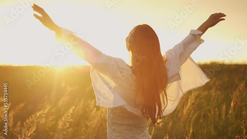 Woman half-turned with flowing hair on a blooming field looks at the sunset. A girl with long hair fluttering in the wind raises her hands up.  photo
