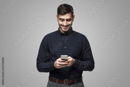 Portrait of cheerful businessman typing sms on smart phone, isolated on gray © Damir Khabirov