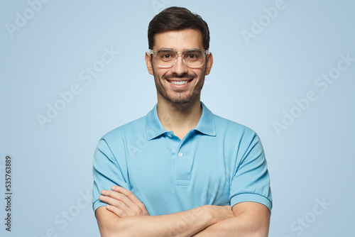 Handsome young guy in polo shirt and trendy glasses standing with crossed arms isolated on blue