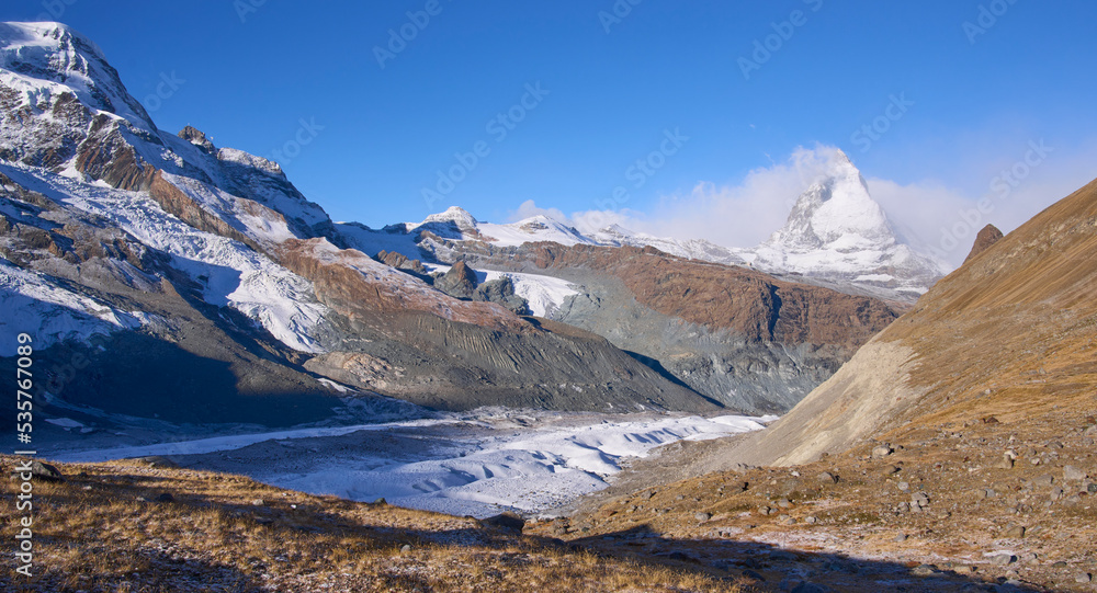 Panoramic view on the iconic mount Matterhorn at background and the Border glacier Grenz Gletscher at foreground in a sunny autumn day, seen from a trail to Monte Rosa hut. Clouds covering the summit.