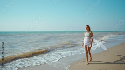 slow motion summer and travel concept - happy smiling young woman in bikini swimsuit with pareo on beach. happy joyful slim fitbody girl walks enjoying wind and waves on sea coast after storm. photo