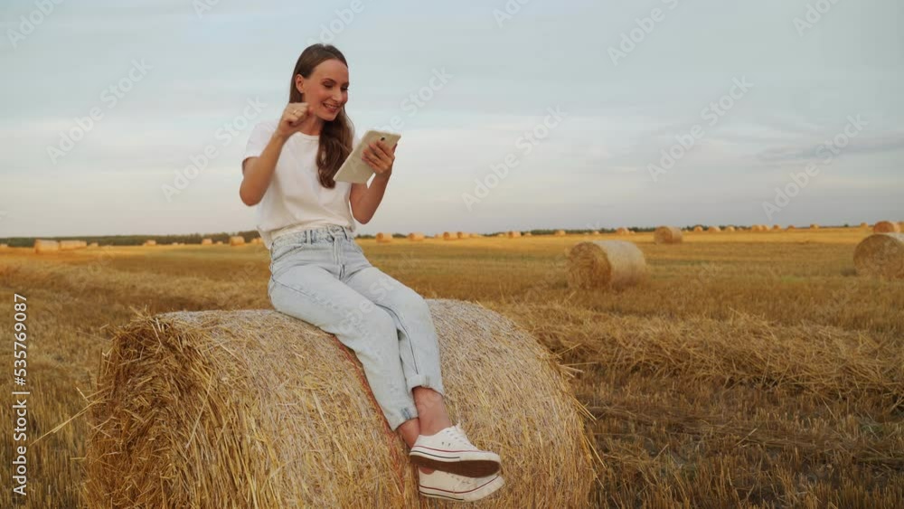 An excited woman reads amazing news on her smartphone while sitting on a haystack in a wheat field at sunset. An excited woman reads amazing news on her smartphone while sitting on a haystack in a wheat field at sunset.
