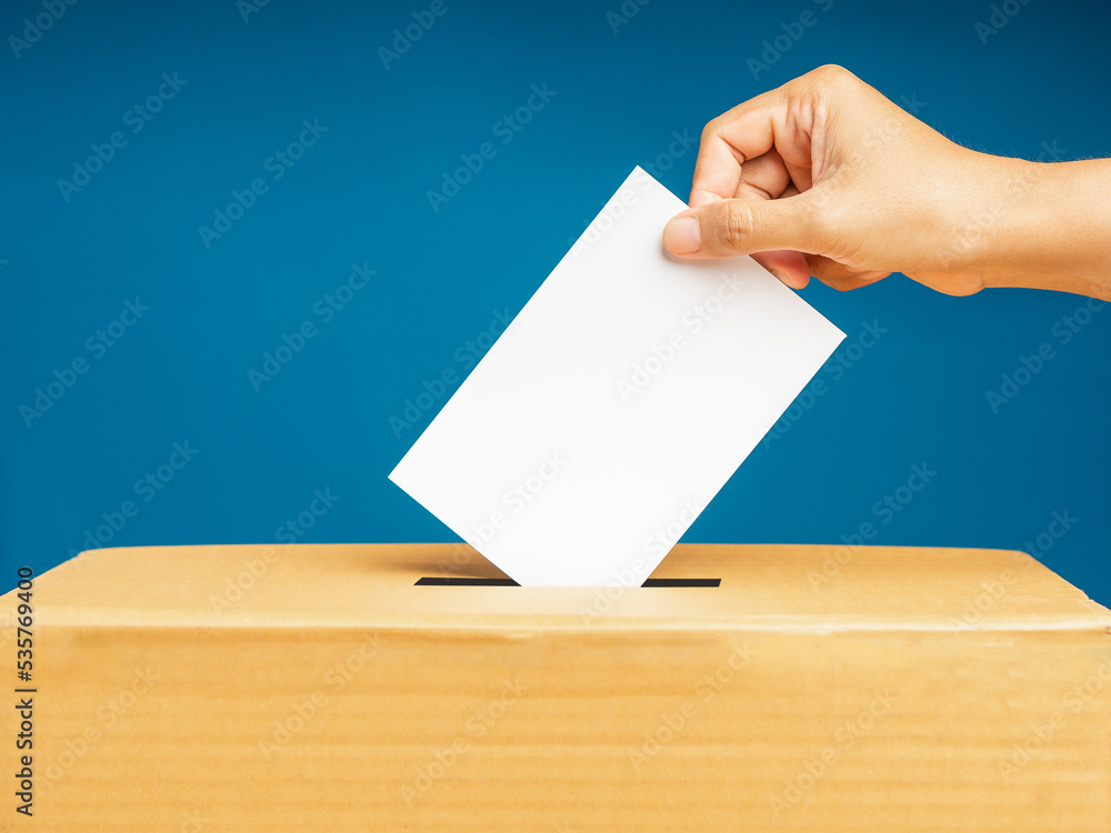Hand voter holding ballot paper putting into the voting box at place election against a blue background Hand voter holding ballot paper putting into the voting box at place election against a blue background
