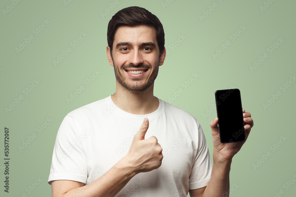 Handsome smiling young man holding blank smartphone, showing thubms up gesture, isolated on green Handsome smiling young man holding blank smartphone, showing thubms up gesture, isolated on green
