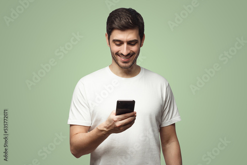 Young man in white t-shirt standing in front of camera, looking with smile at phone screen © Damir Khabirov