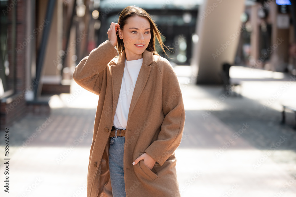 young woman standing with hand in pocket of beige coat and looking away on blurred street.