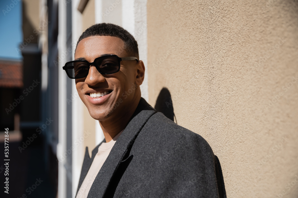 happy african american man in coat and stylish sunglasses looking at camera near wall. happy african american man in coat and stylish sunglasses looking at camera near wall.