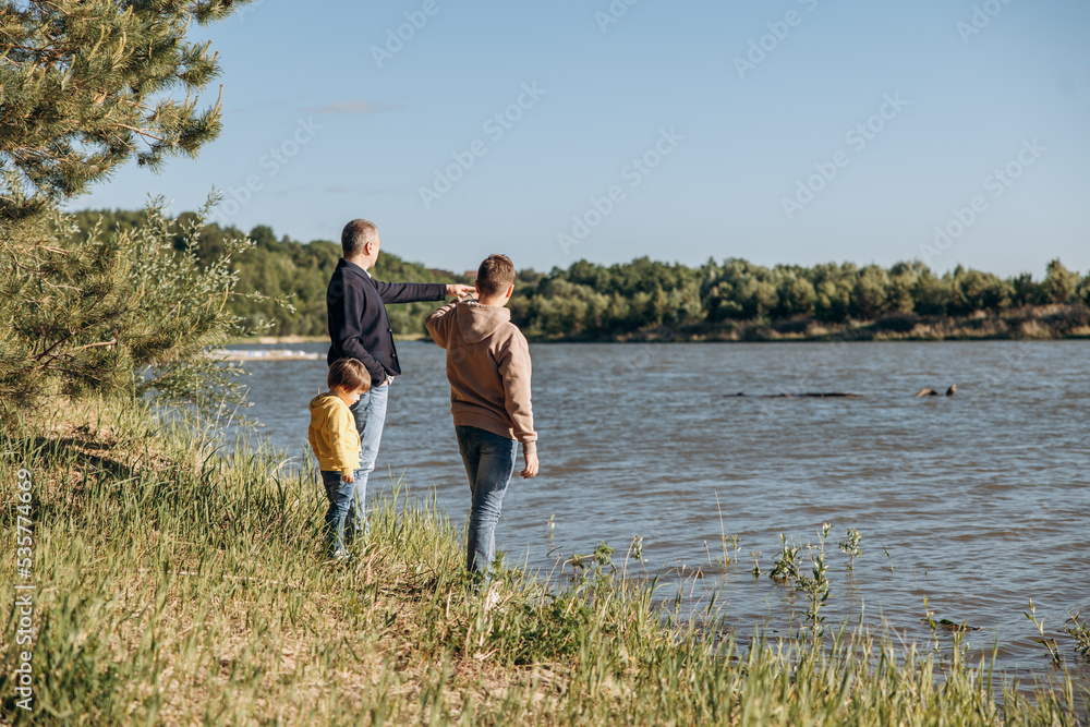 Father and two sons walking along river bank. Family vacation in nature by water. Rear view of dad and children enjoying nature in countryside together. Father and two sons walking along river bank. Family vacation in nature by water. Rear view of dad and children enjoying nature in countryside together.
