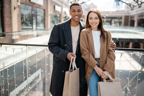 cheerful multiethnic couple in trendy coats holding shopping bags and looking at camera near blurred mall.