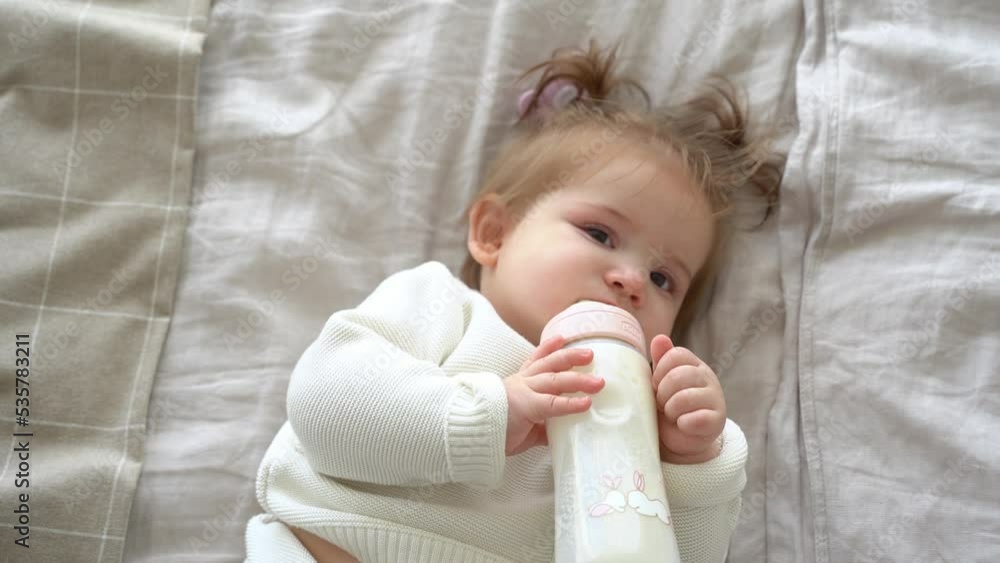 Little baby girl sucking milk from bottle at home, portrait infant eating, drinking powdered milk infant formula. Cute child lying on back on bed, wear knitted sweater, pigtails, look at camera. Little baby girl sucking milk from bottle at home, portrait infant eating, drinking powdered milk infant formula. Cute child lying on back on bed, wear knitted sweater, pigtails, look at camera.