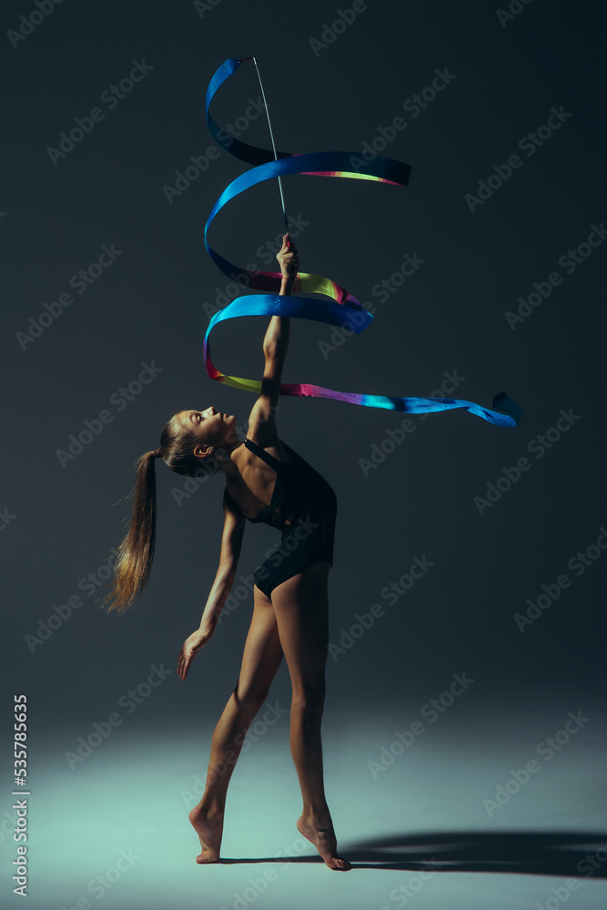 Gymnast child girl lit by light performs gymnastic exercises with ribbon in her hand.
