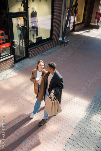 high angle view of interracial couple in trendy coats walking with shopping bags and coffee to go along showcases.
