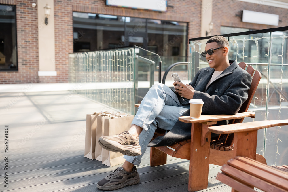 trendy african american man in sunglasses sitting with smartphone near paper cup and shopping bags. trendy african american man in sunglasses sitting with smartphone near paper cup and shopping bags.