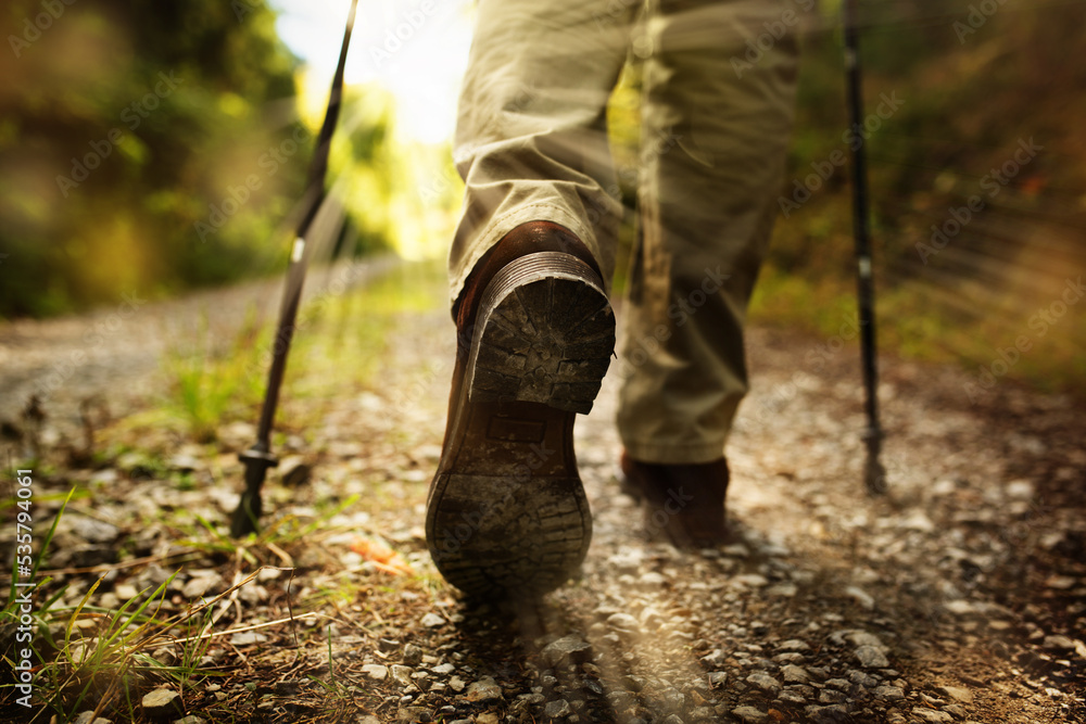 Hiker man with trekking sticks runs on a gravelled dirt trail in the terrain. Horizontal partial shot from behind with short depth of field and focus on boot. Space for text. Hiker man with trekking sticks runs on a gravelled dirt trail in the terrain. Horizontal partial shot from behind with short depth of field and focus on boot. Space for text.
