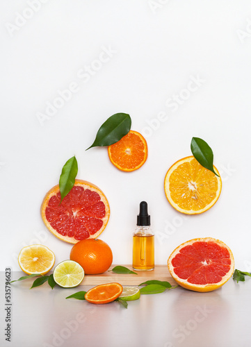 Cosmetic serum with vitamin C and citrus extract on wooden podium and sliced ​​fruits orange, tangerine, lemon and grapefruit on light background. Rejuvenating natural beauty products for facial care