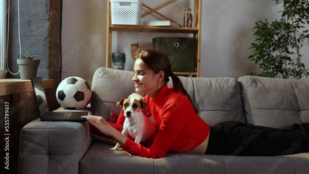 Young woman is using laptop and lying with Jack Russell Terrier dog at home. Beautiful girl hugging with her pet and looking at computer screen with smile. Domestic animal, football fan concept Young woman is using laptop and lying with Jack Russell Terrier dog at home. Beautiful girl hugging with her pet and looking at computer screen with smile. Domestic animal, football fan concept