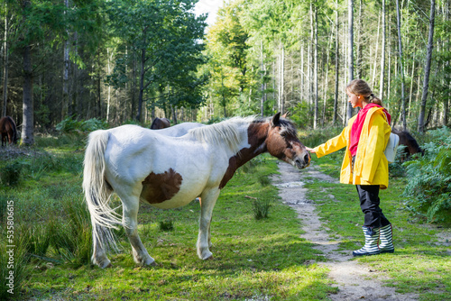 Young smiling woman in a bright yellow raincoat stroking a beautiful wild white horse walking in the forest during the sunny autumn or spring day. Relax on nature. Selective focus.