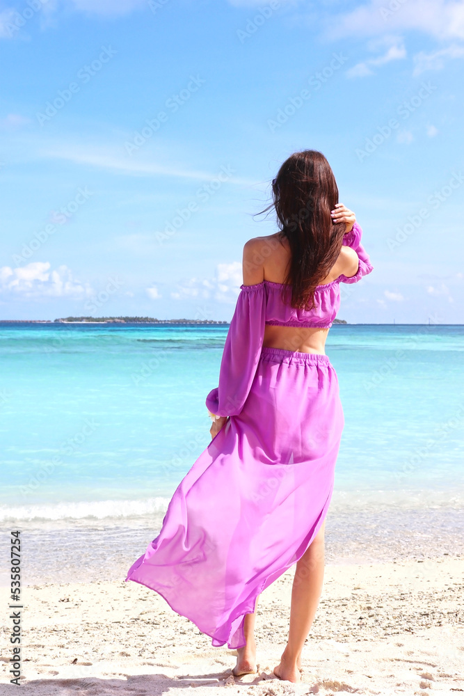 Luxury fashion. Elegant fashion model. Stylish female model in lilac beach cape, standing with her back to the beautiful turquoise ocean in the Maldives Luxury fashion. Elegant fashion model. Stylish female model in lilac beach cape, standing with her back to the beautiful turquoise ocean in the Maldives