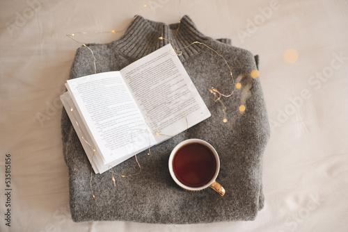 Cup of coffee stay on knitted textile sweater in bed with paper open book with christmas glowing lights close up. Top view.