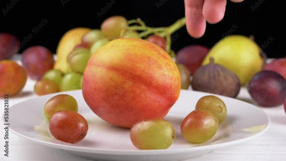 Female Hand Putting a Ripe Apple or Peach on a Plate with Grape and Fruit. Many different colorful fruits, plums, pears lie on the table. Harvest. Black and white background. Variety exotic products. Female Hand Putting a Ripe Apple or Peach on a Plate with Grape and Fruit. Many different colorful fruits, plums, pears lie on the table. Harvest. Black and white background. Variety exotic products.
