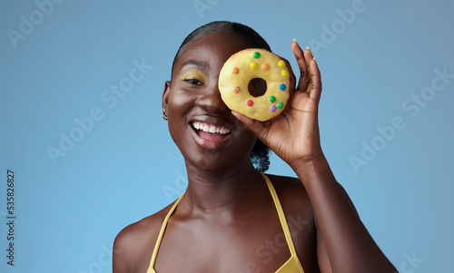 Beauty, black woman and portrait of donut on face with blue studio wall for happy summer style. Matching, beautiful and fun african american girl with yellow makeup for quirky fashion campaign.