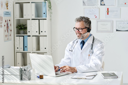 Billede på lærred Mature doctor in headphones sitting at table in front of computer and having con
