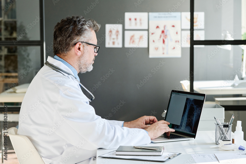 Mature medical professional examining x-ray image on laptop at table at his office
