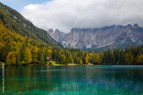 Amazing clear mountain lake in forest among fir trees in sunshine. Bright scenery with beautiful turquoise lake against the background of snow-capped mountains