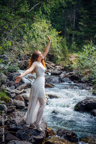 Slender long-haired brunette in white dress posing against small mountain river and green trees. Beautiful young woman walking along the forest stream shore. Vertical image.
