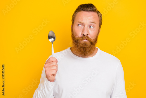 Photo portrait of nice young man suspicious look empty space hold spoon wear stylish white outfit isolated on yellow color background
