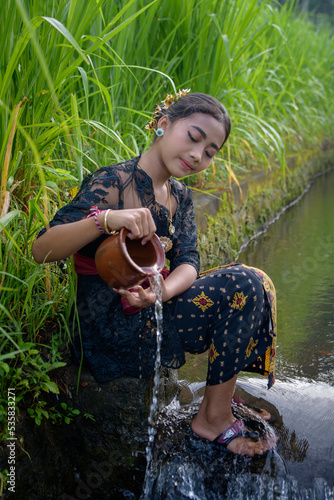 Young Bali girl in traditional clothing sits by the Subak irrigation canal.