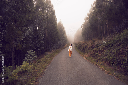 woman walking along a road surrounded by trees with a lot of fog in the background
