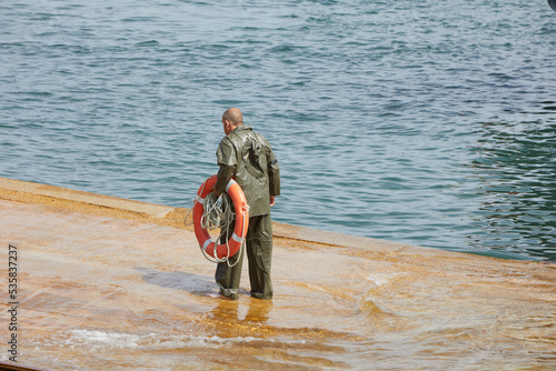 sailor in the port with an orange lifebuoy in his hands. He is dressed in a green waterproof suit.
