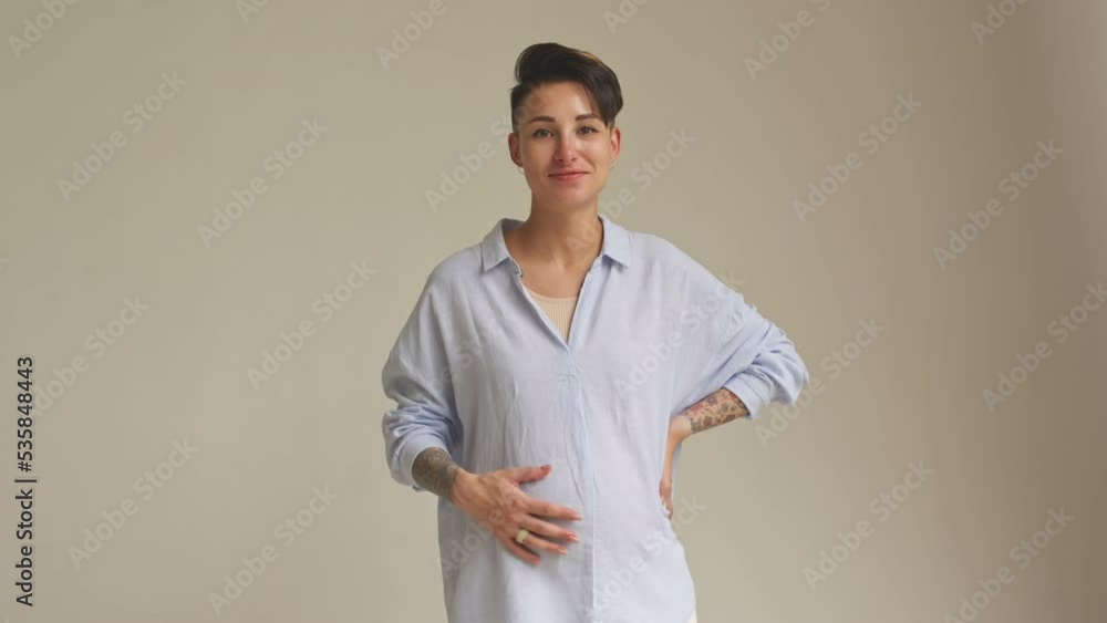 Medium slowmo portrait of young Caucasian pregnant woman with short brown hair and tattoos on hands stroking her belly and smiling at camera standing on clear studio background Medium slowmo portrait of young Caucasian pregnant woman with short brown hair and tattoos on hands stroking her belly and smiling at camera standing on clear studio background