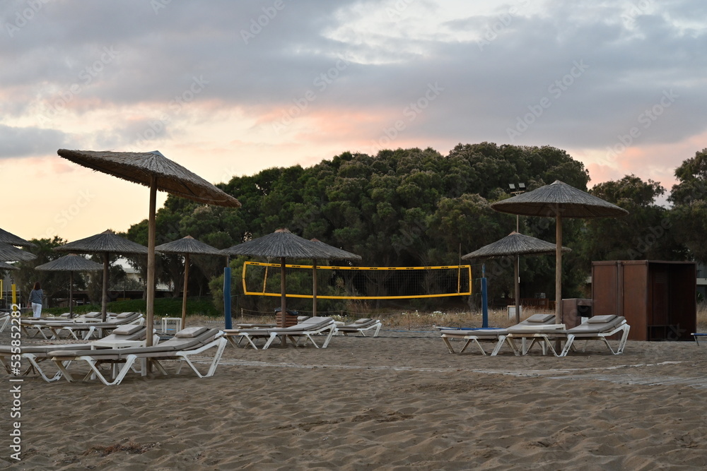 Fototapeta premium Sandy beach of a holiday resort with plastic deck chairs and straw beach umbrellas during sunrise. Behind is playground for beach volleyball with yellow net under clouds.