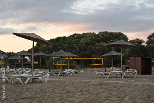 Fototapeta Naklejka Na Ścianę i Meble -  Sandy beach of a holiday resort with plastic deck chairs and straw beach umbrellas during sunrise. Behind is playground for beach volleyball with yellow net under clouds.