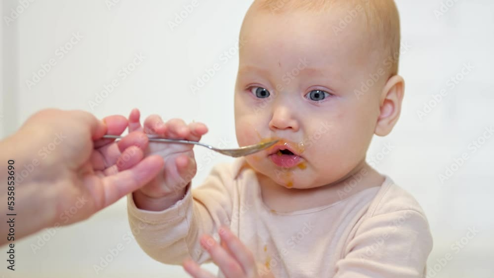 Mother feeds excited baby girl with spoon. Amused daughter tastes sour fruit puree from spoon and grimaces without enjoying flavour in kitchen Mother feeds excited baby girl with spoon. Amused daughter tastes sour fruit puree from spoon and grimaces without enjoying flavour in kitchen