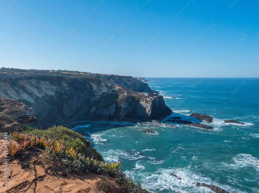 Wild Rota Vicentina coast sea shore with ocean waves and sharp rocks, stones, cliffs and and green vegetation near Almograve, Portugal. Sunny day, blue sky