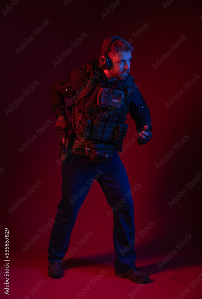 soldier in full gear with weapons. a man in headphones, body armor, with a backpack and a belt. red background. colored, blue-red light