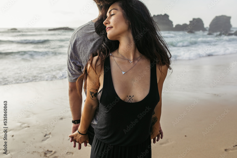 tattooed woman and man standing back to back and holding hands near ocean. tattooed woman and man standing back to back and holding hands near ocean.