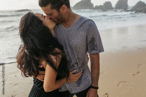 bearded man in grey t-shirt kissing girlfriend near ocean in portugal.
