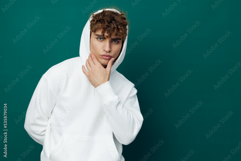 a handsome man stands on a green background in a white hoodie with a hood on his head and looks thoughtfully into the crowd, touching his chin with his hand