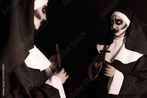 Scary devilish nun with cross near mirror on black background. Halloween party look