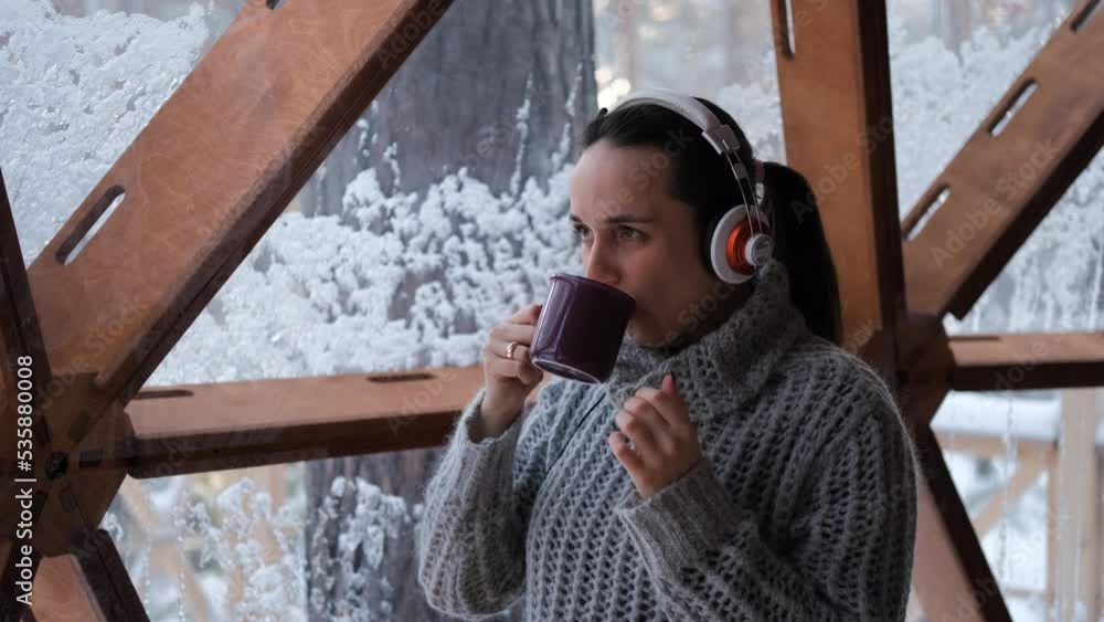 Young happy woman in headphones drinking cocoa or hot chocolate and listening to music while enjoying scenic snowy landscape from dome camping, glamping vacation and winter holidays concept Young happy woman in headphones drinking cocoa or hot chocolate and listening to music while enjoying scenic snowy landscape from dome camping, glamping vacation and winter holidays concept