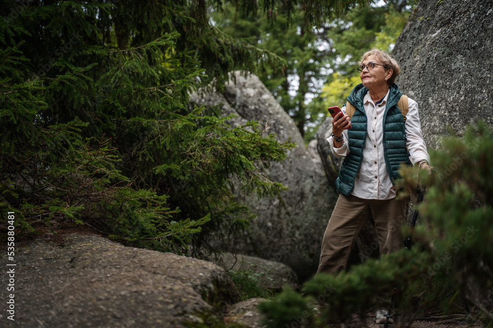 Happy active senior mature woman walking in the forest and using smartphone for navigation. Travel, active retirement and healthy lifestyle concept